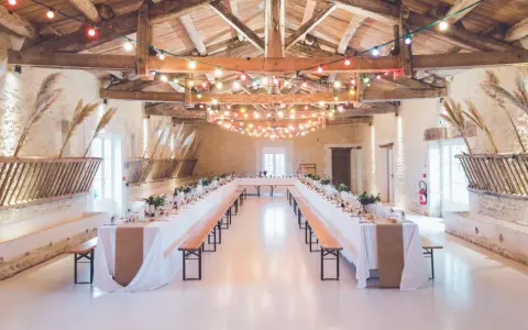 Festlich geschmückte alte Scheune für eine Hochzeit, mit langen Tafeln, Bänken und bunten Lichterketten unter Holzbalken.