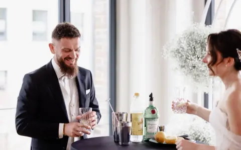 Ein lachender Bräutigam mit einem Drink in der Hand, während die Braut ihm ein Glas reicht. Ein Moment der Freude vor ihrer rituale freie trauung.