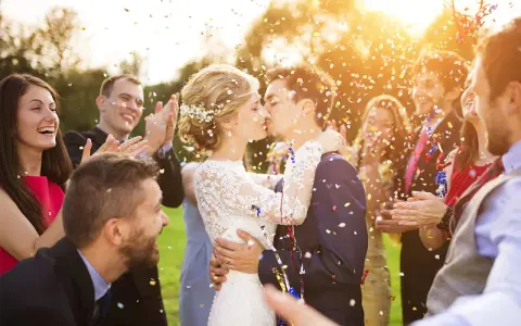 Glückliches Brautpaar küsst sich im Konfettiregen, umgeben von lachenden Gästen. Tolle Spiele zur Hochzeit!