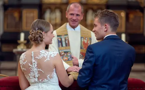 Ein glückliches Paar bei einer christlichen Hochzeit, umgeben von der feierlichen Atmosphäre einer Kirche. Der Bräutigam im blauen Anzug und die Braut im Spitzenkleid blicken sich an, während der Pfarrer sie segnet.
