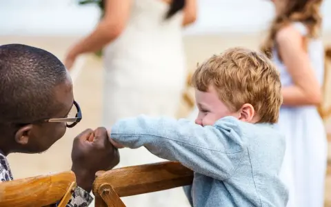 Ein Mann mit Brille hält die Hand eines Jungen. Im Hintergrund heiratet mein Sohn.
