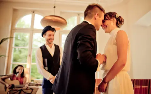 Ein Brautpaar teilt einen innigen Moment, während ein Mann mit Hut und eine Frau mit Gitarre im Hintergrund stehen. Die emotionale Rede beim Standesamt wird durch ihre Freude spürbar.