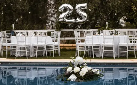 Festlich gedeckte Tische mit weißen Stühlen und Luftballons "25" über ihnen – perfekt für eine Rede zur Silberhochzeit am Pool.