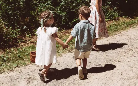 Zwei Kinder, ein Mädchen mit Blumenkranz und ein Junge, halten Händchen auf dem Weg zu einer Hochzeit mit Kindern.