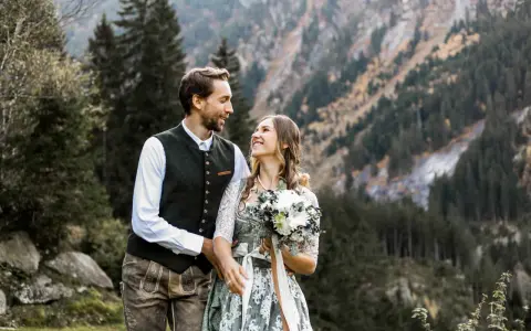 Ein glückliches Brautpaar feiert seine Hochzeit auf der Alm. Sie tragen traditionelle Tracht vor einer Bergkulisse.