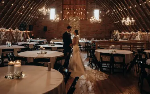 Ein Brautpaar steht sich in einem rustikalen Saal gegenüber, der für eine minimalistische Hochzeit mit Lichterketten und Kerzen dekoriert ist.