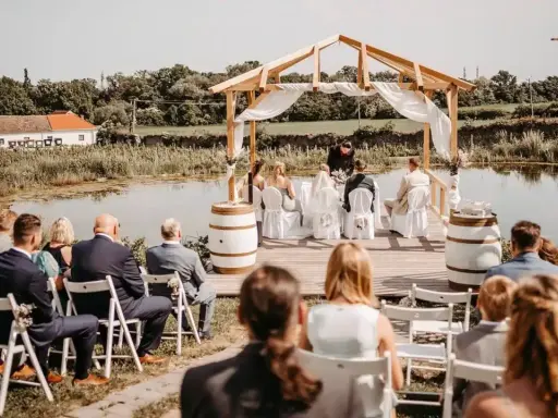 Eine romantische **Hochzeit im Freien** am See. Gäste sitzen auf Stühlen, das Brautpaar steht unter einem Holzdach.