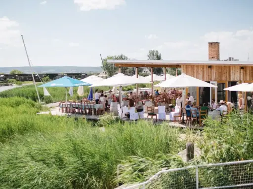 Gäste genießen eine festliche Hochzeit am Wasser. Sonnenschirme spenden Schatten, Tische sind für das Festessen gedeckt.