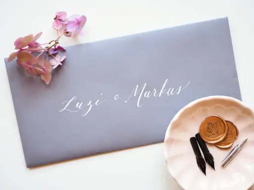 Grauer Umschlag mit weißer Kalligrafie "Luzi & Markus". Daneben eine rosa Schale mit Siegelwachs, Stiften und Federn. Was schreibt man auf den Umschlag zur Hochzeit?