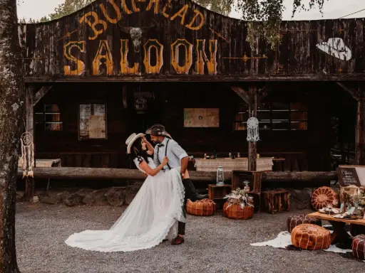 Ein Brautpaar feiert seine western hochzeit vor einem Saloon. Sie tanzen ausgelassen, umgeben von rustikaler Deko.
