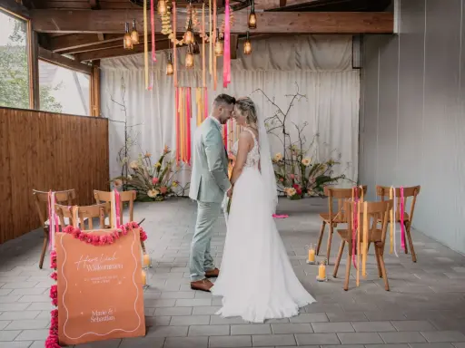 Brautpaar im boho style wedding, umgeben von Blumen und Bändern. Ein Willkommensschild steht im Vordergrund.