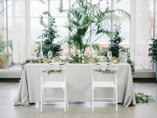 Ein festlich gedeckter Tisch für eine greenery hochzeit, umgeben von üppigem Grün. Zwei weiße Stühle mit Eukalyptus-Girlanden stehen bereit.