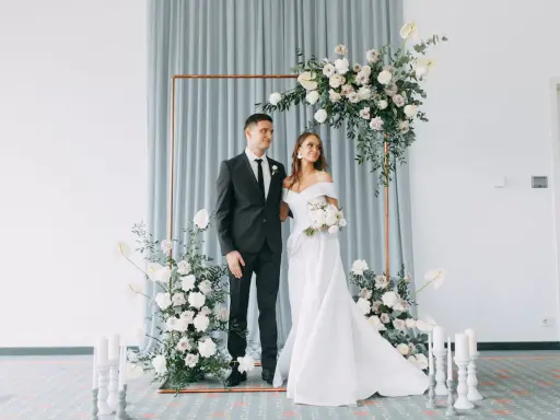 Ein Brautpaar steht vor einem floralen Bogen, bereit, wie heiratet man. Sie strahlen Glück aus.