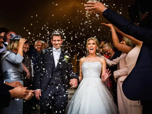 Glückliches Brautpaar bei einer englischen Hochzeit, umgeben von Blütenblättern und jubelnden Gästen.