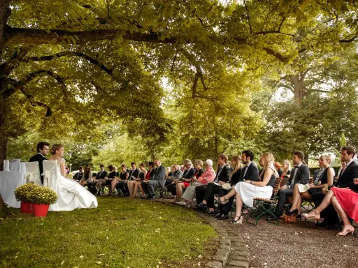 Hochzeit im Freien unter einem Baum. Gäste sitzen auf Bänken. Tolle Ideen zur Hochzeit für eine romantische Zeremonie.
