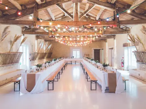 Festlich geschmückte alte Scheune für eine Hochzeit, mit langen Tafeln, Bänken und bunten Lichterketten unter Holzbalken.