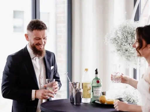 Ein lachender Bräutigam mit einem Drink in der Hand, während die Braut ihm ein Glas reicht. Ein Moment der Freude vor ihrer rituale freie trauung.