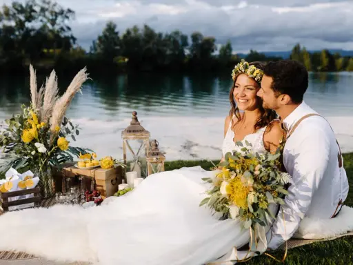 Glückliches Brautpaar genießt seine Hochzeit am See. Blumen, Laternen und ein Picknickkorb schaffen romantische Atmosphäre.