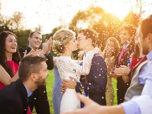 Glückliches Brautpaar küsst sich im Konfettiregen, umgeben von lachenden Gästen. Tolle Spiele zur Hochzeit!