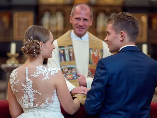 Ein glückliches Paar bei einer christlichen Hochzeit, umgeben von der feierlichen Atmosphäre einer Kirche. Der Bräutigam im blauen Anzug und die Braut im Spitzenkleid blicken sich an, während der Pfarrer sie segnet.