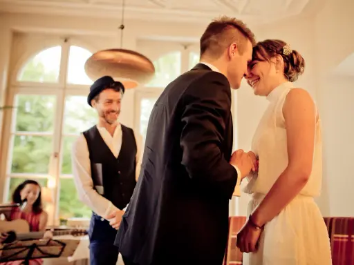 Ein Brautpaar teilt einen innigen Moment, während ein Mann mit Hut und eine Frau mit Gitarre im Hintergrund stehen. Die emotionale Rede beim Standesamt wird durch ihre Freude spürbar.