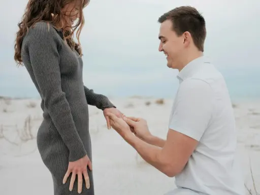 Ein Mann kniet am Strand und steckt seiner lachenden Partnerin einen Ring an den Finger. Ein Moment, der die tiefe **Verlobung Bedeutung** symbolisiert.