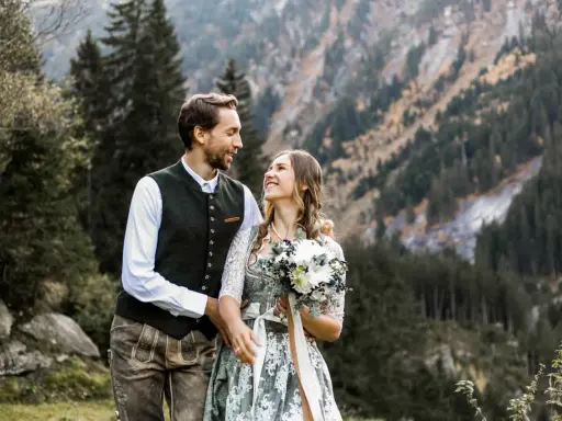 Ein glückliches Brautpaar feiert seine Hochzeit auf der Alm. Sie tragen traditionelle Tracht vor einer Bergkulisse.
