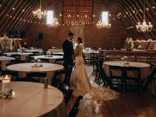 Ein Brautpaar steht sich in einem rustikalen Saal gegenüber, der für eine minimalistische Hochzeit mit Lichterketten und Kerzen dekoriert ist.