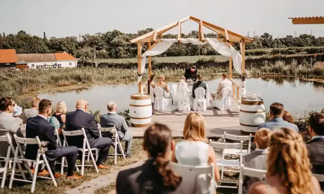 Eine romantische **Hochzeit im Freien** am See. Gäste sitzen auf Stühlen, das Brautpaar steht unter einem Holzdach.