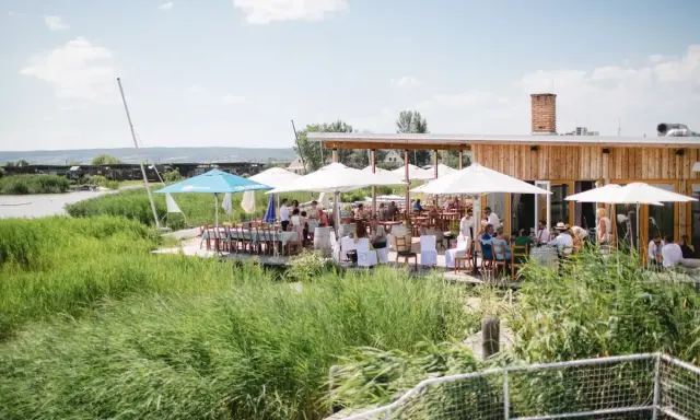 Gäste genießen eine festliche Hochzeit am Wasser. Sonnenschirme spenden Schatten, Tische sind für das Festessen gedeckt.