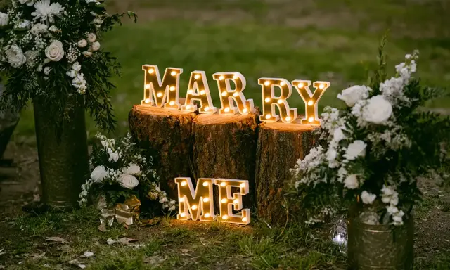 Leuchtende Buchstaben "MARRY ME" auf Baumstämmen, umgeben von Blumen. Perfekte heiratsantrag ideen für einen romantischen Moment.