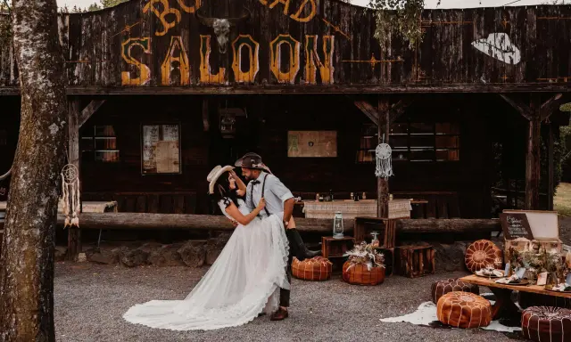 Ein Brautpaar feiert seine western hochzeit vor einem Saloon. Sie tanzen ausgelassen, umgeben von rustikaler Deko.