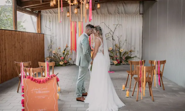 Brautpaar im boho style wedding, umgeben von Blumen und Bändern. Ein Willkommensschild steht im Vordergrund.
