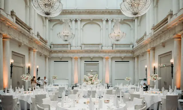 Elegante Tische mit weißen Hussen und Blumenarrangements in einem prunkvollen Saal mit Kronleuchtern – eine besondere Hochzeitslocation.