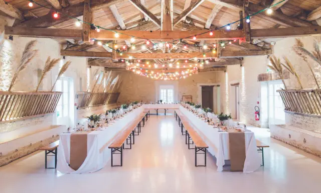 Festlich geschmückte alte Scheune für eine Hochzeit, mit langen Tafeln, Bänken und bunten Lichterketten unter Holzbalken.