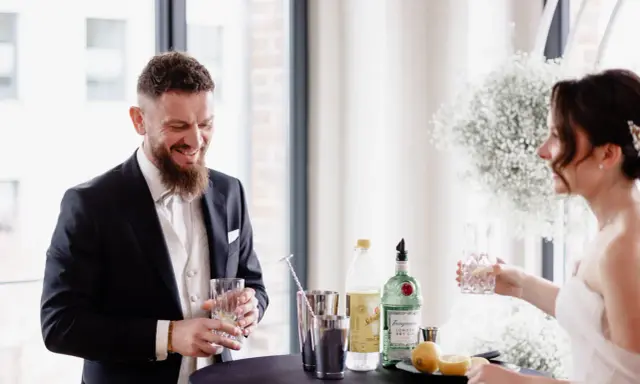 Ein lachender Bräutigam mit einem Drink in der Hand, während die Braut ihm ein Glas reicht. Ein Moment der Freude vor ihrer rituale freie trauung.