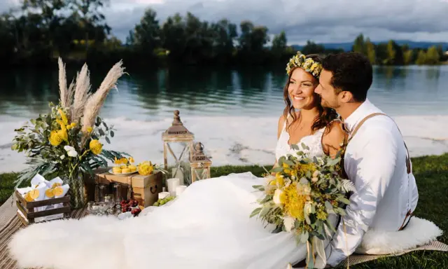 Glückliches Brautpaar genießt seine Hochzeit am See. Blumen, Laternen und ein Picknickkorb schaffen romantische Atmosphäre.
