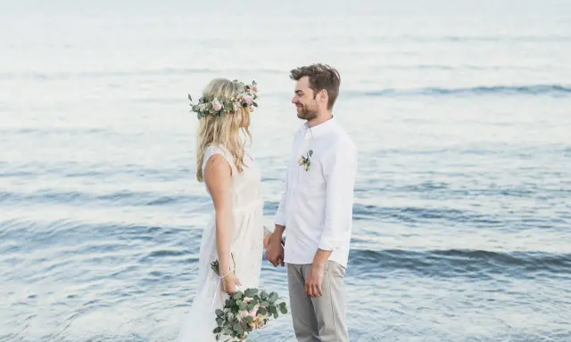 Ein glückliches Paar feiert seine Hochzeit am Strand zu zweit. Die Braut trägt einen Blumenkranz, der Bräutigam ein weißes Hemd.