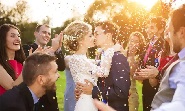 Glückliches Brautpaar küsst sich im Konfettiregen, umgeben von lachenden Gästen. Tolle Spiele zur Hochzeit!