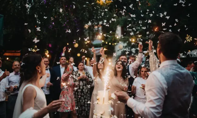 Eine ausgelassene Hochzeitsparty statt einer steifen Feier! Gäste werfen Schmetterlinge und zünden Wunderkerzen an, während das Brautpaar vor einem beleuchteten Kuchen steht.
