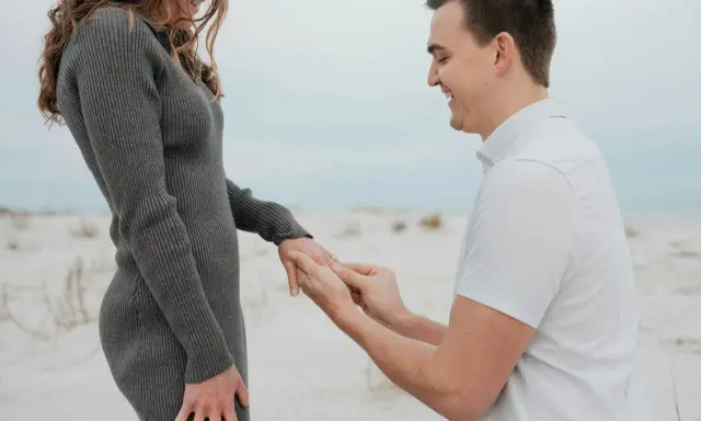 Ein Mann kniet am Strand und steckt seiner lachenden Partnerin einen Ring an den Finger. Ein Moment, der die tiefe **Verlobung Bedeutung** symbolisiert.