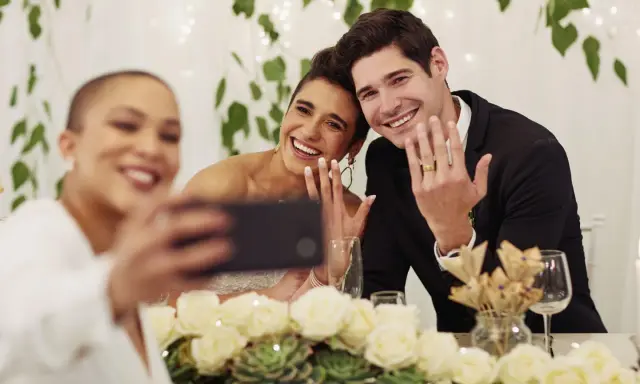 Brautpaar zeigt Ringe für ein Hochzeitsfoto, während eine Freundin ein Selfie macht.