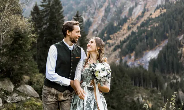 Ein glückliches Brautpaar feiert seine Hochzeit auf der Alm. Sie tragen traditionelle Tracht vor einer Bergkulisse.