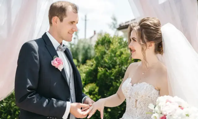 Ein Bräutigam steckt seiner Braut den Ring an. Ein Moment, der ihr Ehe Versprechen besiegelt, unter einem Baldachin aus zartem Stoff.
