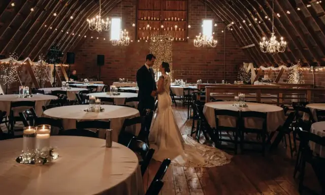 Ein Brautpaar steht sich in einem rustikalen Saal gegenüber, der für eine minimalistische Hochzeit mit Lichterketten und Kerzen dekoriert ist.