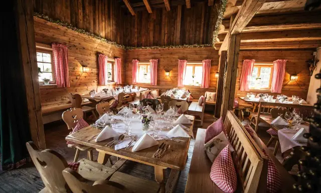Gemütliche Holzhütte, festlich gedeckte Tische für eine Egerner Alm Hochzeit. Rote Karovorhänge und Blumen schmücken den rustikalen Raum.