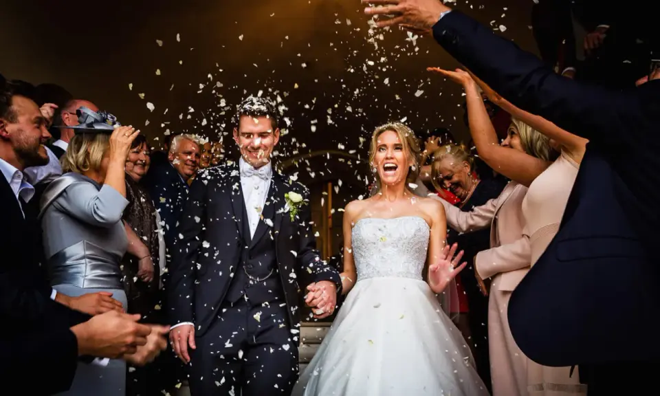 Glückliches Brautpaar bei einer englischen Hochzeit, umgeben von Blütenblättern und jubelnden Gästen.