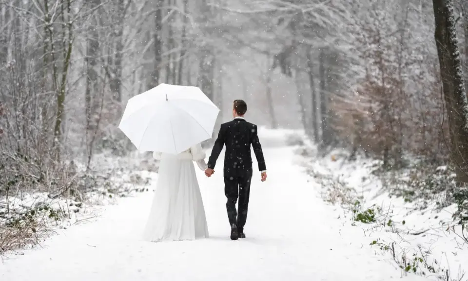 Ein Brautpaar geht Hand in Hand unter einem weißen Schirm durch einen verschneiten Wald. Vielleicht ist dies der beste Monat zum Heiraten für alle, die eine romantische Winterhochzeit wünschen.