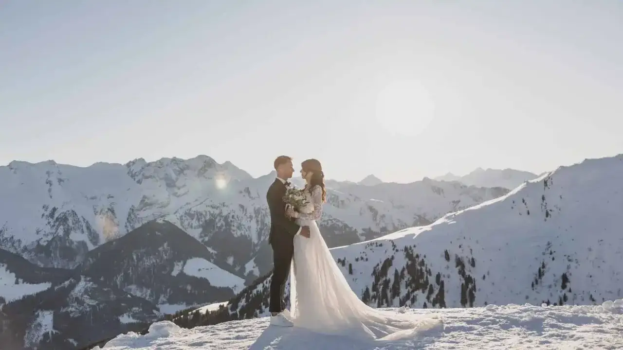 Ein Brautpaar steht sich in der verschneiten Berglandschaft gegenüber. Ihre **Hochzeit im Schnee** ist ein magischer Moment vor der Kulisse schneebedeckter Gipfel.