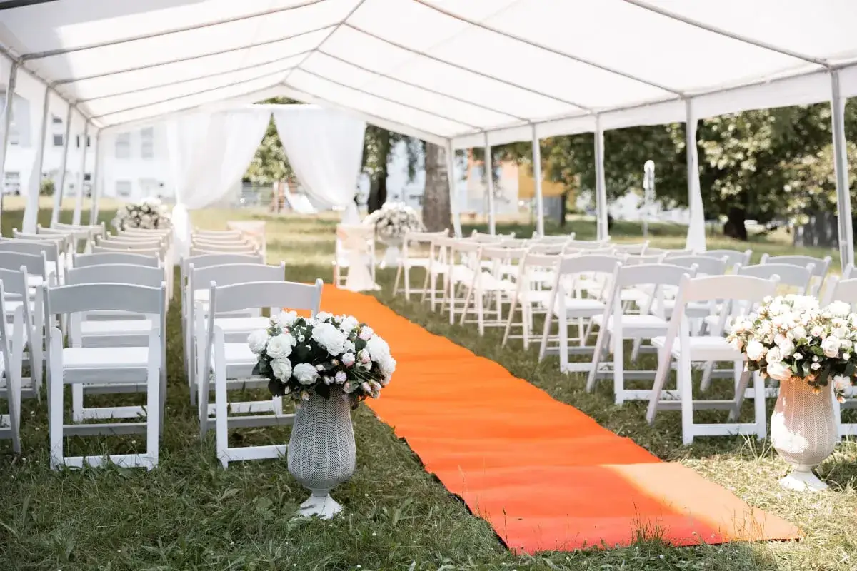 Festliche Hochzeit unter Zelt: weiße Stühle, orange Teppich und Blumenarrangements.